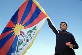Ein schwarz gekleideter Mann schwenkt die Flagge Tibets. Auf der Mitte der Falgge ist eine Sonne; von ihr gehen sechs rote und sechs blaue Strahlen ab; vor der Sonne ist ein schneebedeckter Berg symbolisch dargestellt; davor sind zwei Seelöwen und ein symbolisches Juwel dargestellt. Die gesamte Flagge ist goldgelb umrandet.