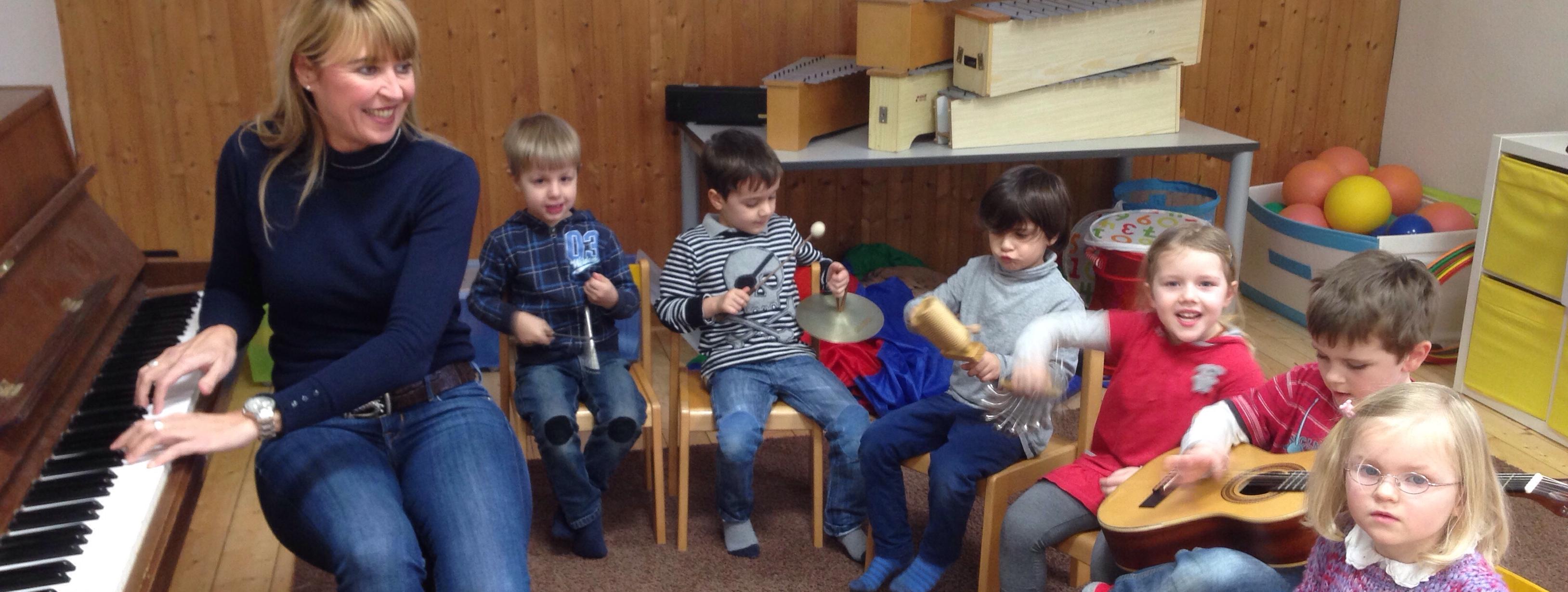 sechs kleine Kinder sitzen auf Stühlen in einem Halbkreis um ihre Musiklehrerin herum. Die Lehrerin hat die Hände auf die Tasten eines Klaviers gelegt und erklärt den Kindern etwas.