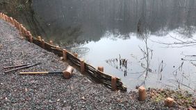 Ein Weg aus grobem Schotter am Hexenteich, knapp vor der Wasserlinie wurden Pflöcke in den Boden getrieben und dazwischen ein Geflächt aus biegsamen Stöcken gewunden. Ein großer Holzhammer liegt auf dem Weg.