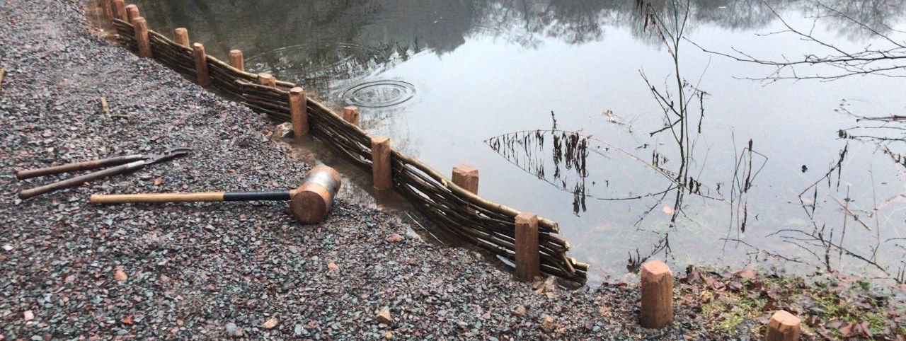 Ein Weg aus grobem Schotter am Hexenteich, knapp vor der Wasserlinie wurden Pflöcke in den Boden getrieben und dazwischen ein Geflächt aus biegsamen Stöcken gewunden. Ein großer Holzhammer liegt auf dem Weg.