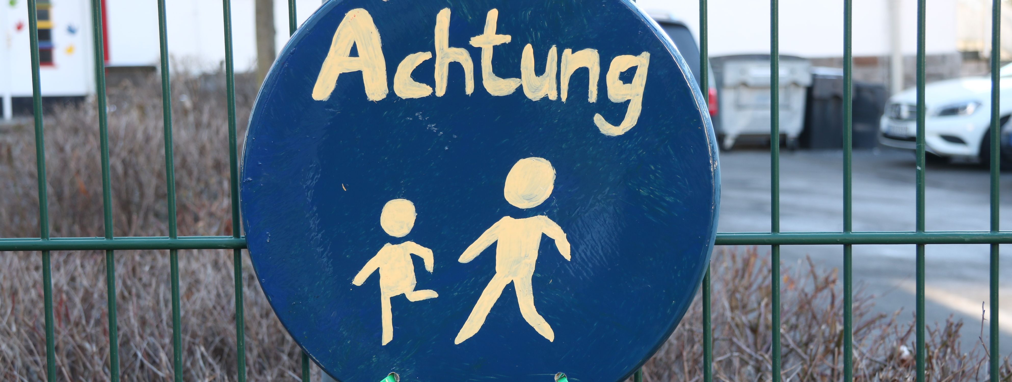 An einem grünen Metallzaun vor der Josefschule hängt ein selbstgemaltes Schild. Es ist rund, darauf sind zwei laufende Strichmännchen in weißer Farbe, darüber steht das Wort "Achtung".