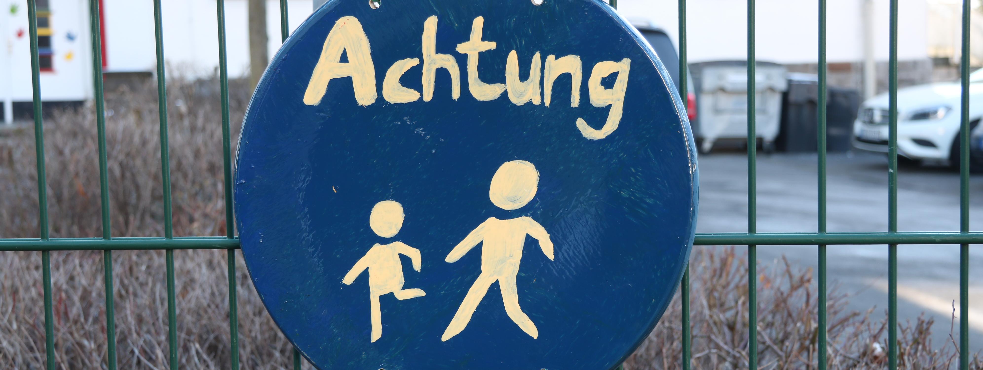 An einem grünen Metallzaun vor der Josefschule hängt ein selbstgemaltes Schild. Es ist rund, darauf sind zwei laufende Strichmännchen in weißer Farbe, darüber steht das Wort "Achtung".
