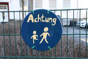 An einem grünen Metallzaun vor der Josefschule hängt ein selbstgemaltes Schild. Es ist rund, darauf sind zwei laufende Strichmännchen in weißer Farbe, darüber steht das Wort "Achtung".