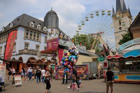 Buntes Kirmestreiben vor dem Alten Rathaus. Viele Passanten schlendern an den Buden und Fahrgeschäften vorbei, in der Mitte steht ein Lufballonverkäufer. Im Hintergrund ist das Riesenrad und der Turm der St. Vincenz-Kirche zu sehen.