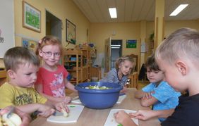 Fünf Kita-Kinder sitzen an einem Tisch und schneiden Äpfel in kleine Stückchen. Die geschnittenen Apfelstücke wandern dann in eine große blaue Schüssel, die mittig auf dem Tisch steht.