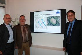 Sebastian Arlt (1. Beigeordneter der Stadt), Frank Wagenbach (Fachbereichsleiter Umwelt, Planen und Bauen) und Steffen Krippenstapel (Geschäftsführer GEWOGE) stehen neben einem Monitor der an einer Wand hängt. Darauf ist der Plan für das neue Wohnbaugebiet zu sehen und eine Luftaufnahme des Sportplatzes, so wie er jetzt noch aussieht.