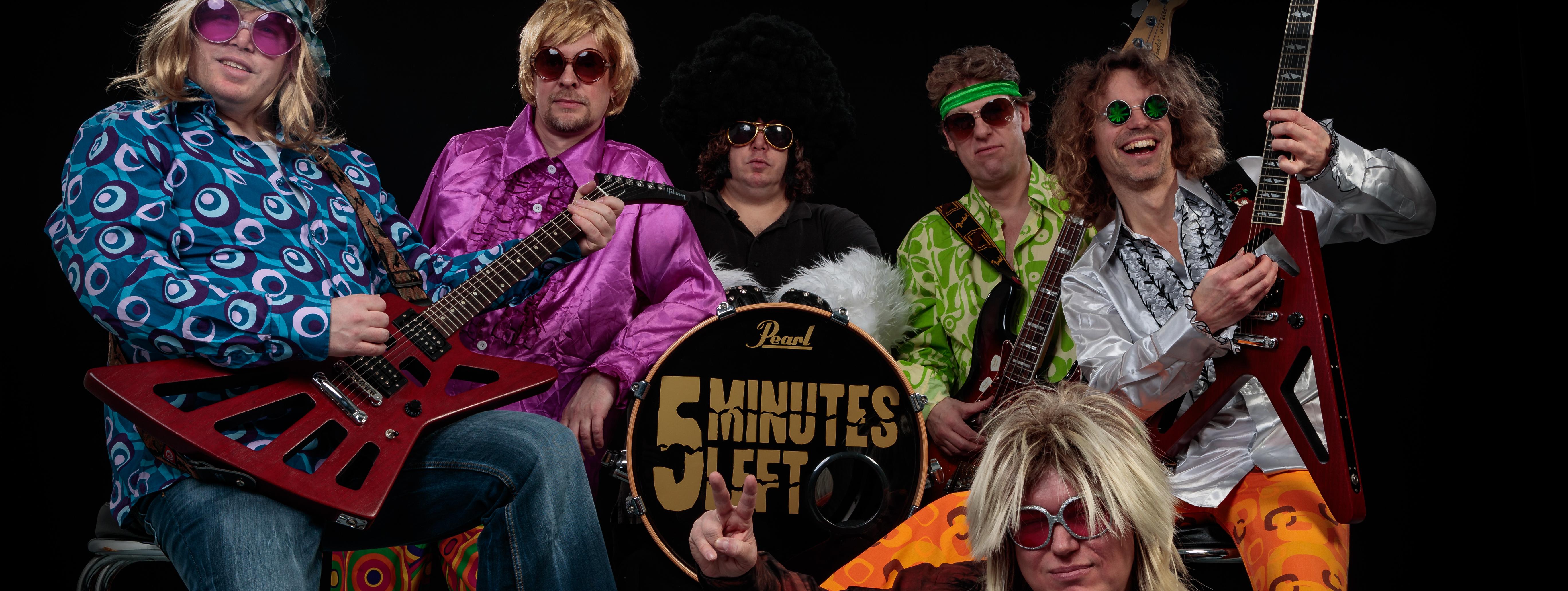 Fünf Musiker sitzen, einer liegt vor ihnen, drei haben E-Gitarren, der Musiker in der Mitte hat die Basedrum seines Schlagzeuges auf dem Schoß. Alle tragen große Sonnenbrillen und schrill-bunte Kleidung aus den 70er Jahren.