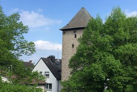 Ein Foto vom Poenigeturm, ein heller geschlämmter viereckiger Steinturm mit dunklem Spitzdach. Abgebildet ist die obere Hälfte des Turmes, an der rechten Seite wird der Turm durch einen Kastanienbaum in vollem Blattgrün verdeckt.