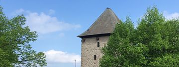 Ein Foto vom Poenigeturm, ein heller geschlämmter viereckiger Steinturm mit dunklem Spitzdach. Abgebildet ist die obere Hälfte des Turmes, an der rechten Seite wird der Turm durch einen Kastanienbaum in vollem Blattgrün verdeckt.