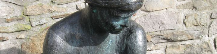 Eine Bronzeskulptur. Sie stellt eine nackte, sitzende Frau dar, deren Blick nachdenktlich nach unten gerichtet ist. Sie sitzt auf der Bank beim Alten Rathaus.