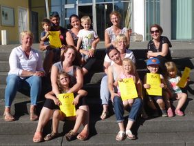 Eltern und Kindersitzen auf Steinstufen, viele halten einen gelben Zettel in der Hand.