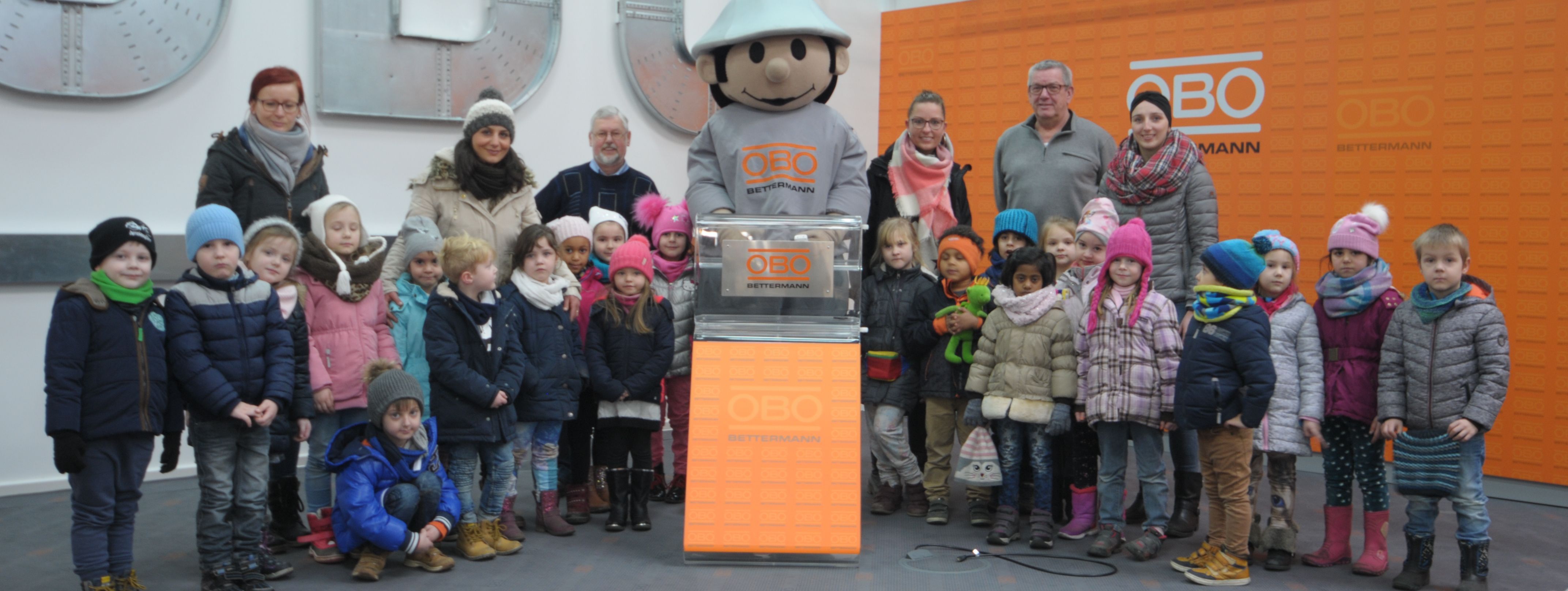 Die Kinder und ihre Erzieherinnen haben sich um das OBO-Maskottchen versammelt. Das Maskottchen ist ein Männchen mit grauem Sicherheitshelm und einem T-Shirt mit dem OBO-Schriftzug. Im Vordergrund sitzen lauter kleine Maskottchen aus Plüsch, die die Kinder als Geschenk mitnehmen durften.