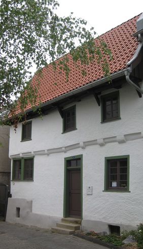Die weiße Außenfassade des Schmarotzerhauses ist zu sehen. Oben im rechten Bildabschnitt ragen die Äste eines Baumes ins Bild. Das Dach des Schmarotzerhauses besteht aus roten Pfannen. 