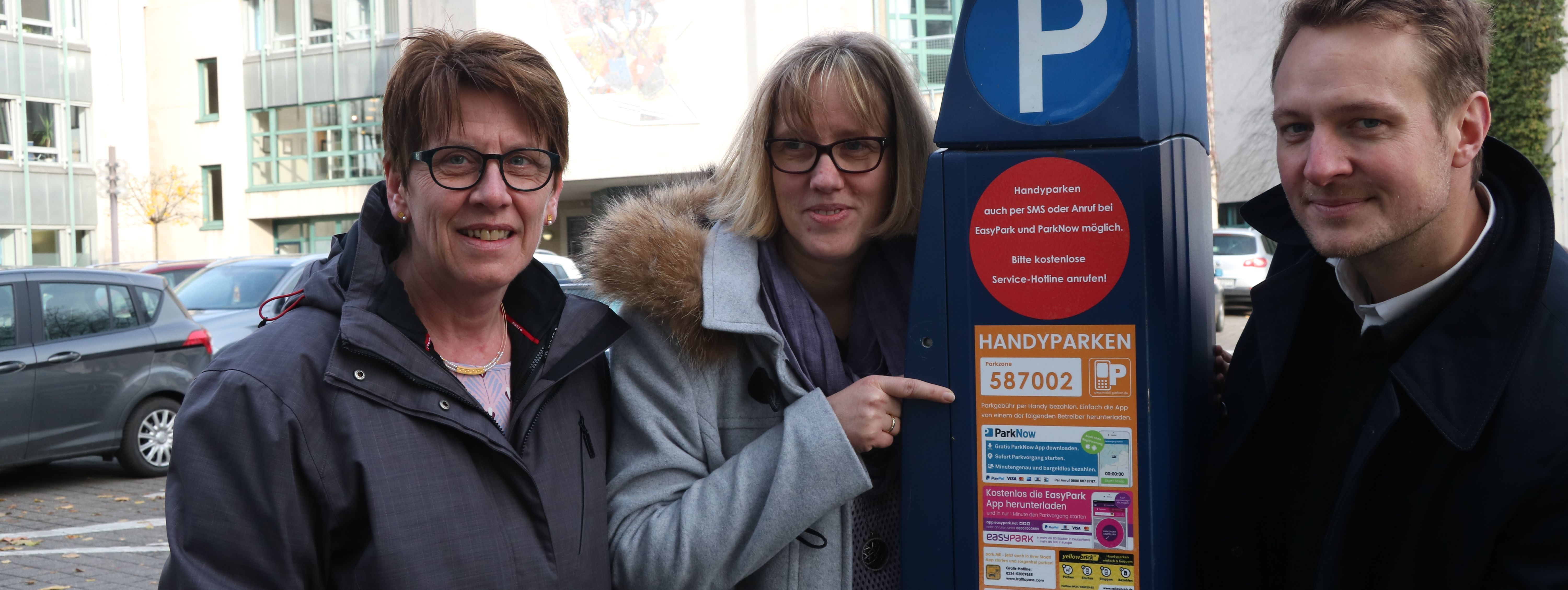 Martina Potthoff, Birgit Sturzenhecker und Philipp Zimmermann stehen neben einem blauen Parkautomaten. Frau Sturzenhecker zeigt mit dem Finger auf den neuen Aufkleber auf dem Automaten, der das Handyparken erklärt.