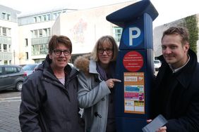 Martina Potthoff, Birgit Sturzenhecker und Philipp Zimmermann stehen neben einem blauen Parkautomaten. Frau Sturzenhecker zeigt mit dem Finger auf den neuen Aufkleber auf dem Automaten, der das Handyparken erklärt.