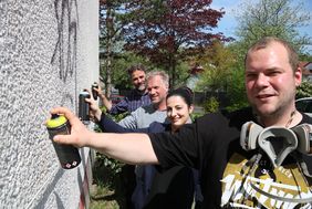 drei Männer und eine Frau stehen an einer Wand und halten Farbspraydosen, so als wollten sie sofort loslegen und ein Grafitti an die Wand sprühen.