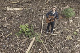 Sem Seiffert steht mit seiner Gitarre mitten im Wald, um ihn herum liegen zahllose abgebrochene Äste