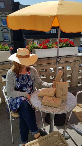 Auf dem Balkon der Stadtbücherei sitzt eine Frau mit einem großen Strohhut, der ihr Gesicht verdeckt. Auf dem Tisch vor ihr liegen in braunem Packpapier verpackte Bücher.