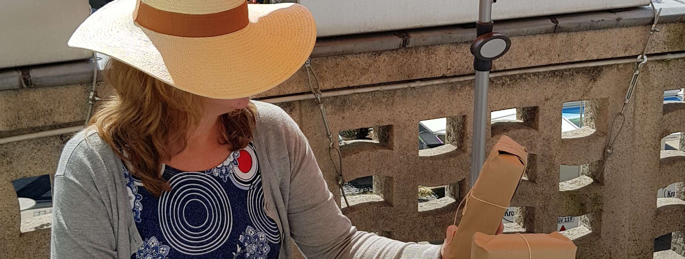 Auf dem Balkon der Stadtbücherei sitzt eine Frau mit einem großen Strohhut, der ihr Gesicht verdeckt. Auf dem Tisch vor ihr liegen in braunem Packpapier verpackte Bücher.