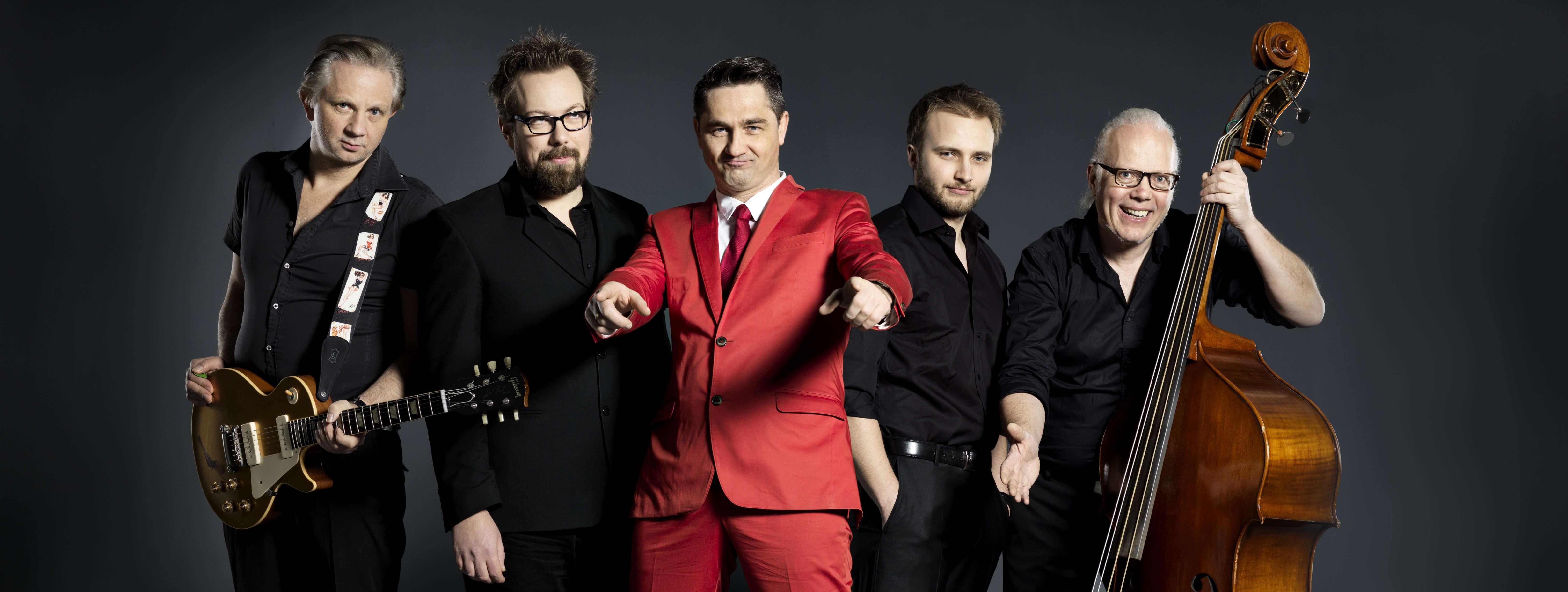 fünf Musiker stehen nebeneinander, alle sind in schwarzen Anzügen gekleides, nur der Sänger in der Mitte trägt rot. Sie halten Instrummente wie Saxophon und Kontrabass