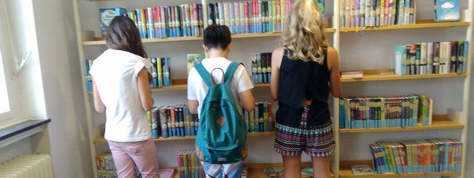 drei Jugendliche stehen vor einem Bücherregal in der Stadtbücherei und suchen sich Lesestoff für die Sommerferien aus