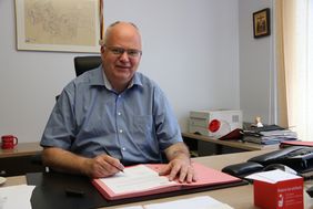 Bürgermeister Martin Wächter sitzt an seinem Schreibtisch, vor ihm liegt eine Briefmappe. In der Hand hält er einen Stift, mit dem er gerade den Vertrag unterschreibt.