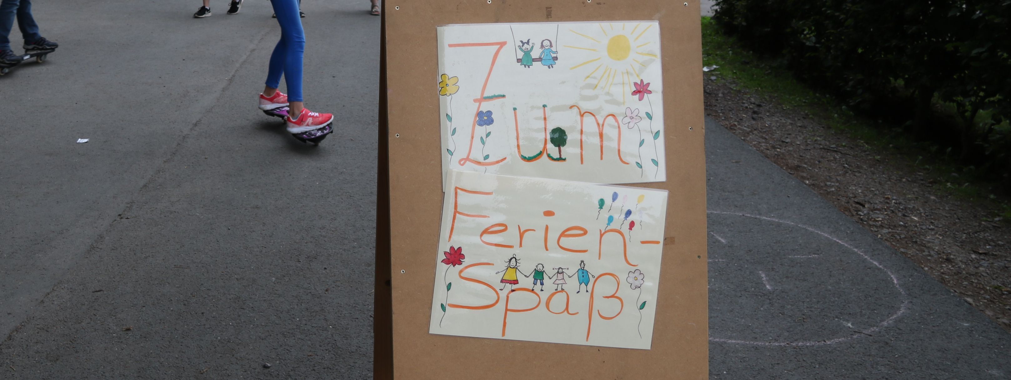 Kinder spielen vor der Jugendbildungsstätte Kluse. Im Vordergrund steht ein Holzschild, das auf den Ferienspaß hinweißt.