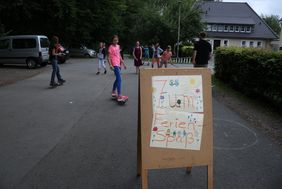 Kinder spielen vor der Jugendbildungsstätte Kluse. Im Vordergrund steht ein Holzschild, das auf den Ferienspaß hinweißt.