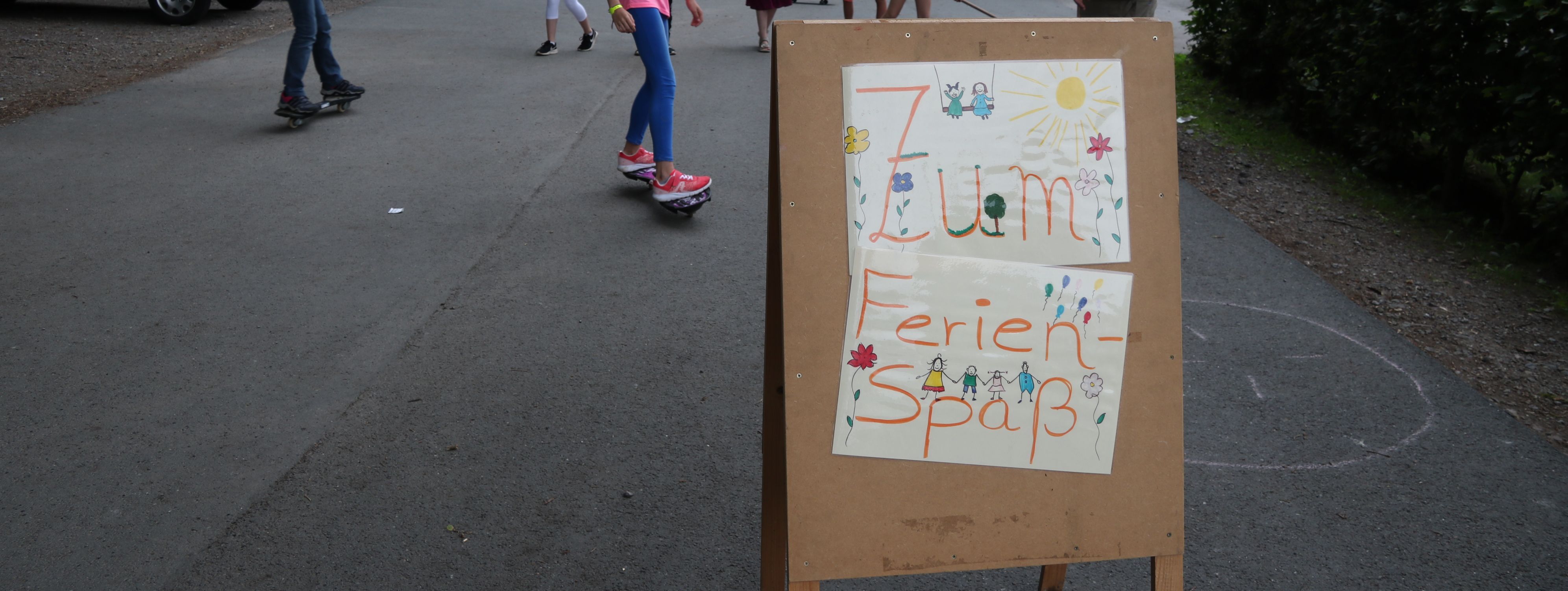 Kinder spielen vor der Jugendbildungsstätte Kluse. Im Vordergrund steht ein Holzschild, das auf den Ferienspaß hinweißt.