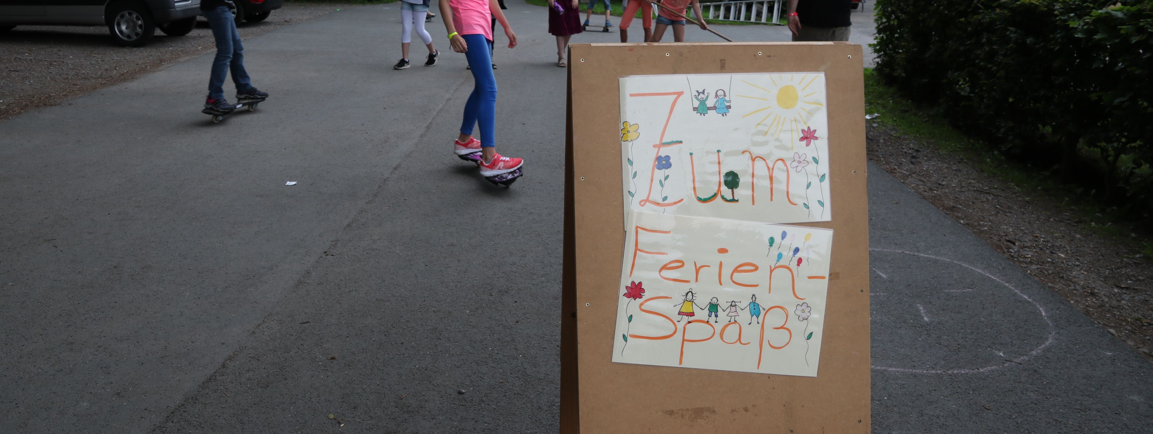 Kinder spielen vor der Jugendbildungsstätte Kluse. Im Vordergrund steht ein Holzschild, das auf den Ferienspaß hinweißt.