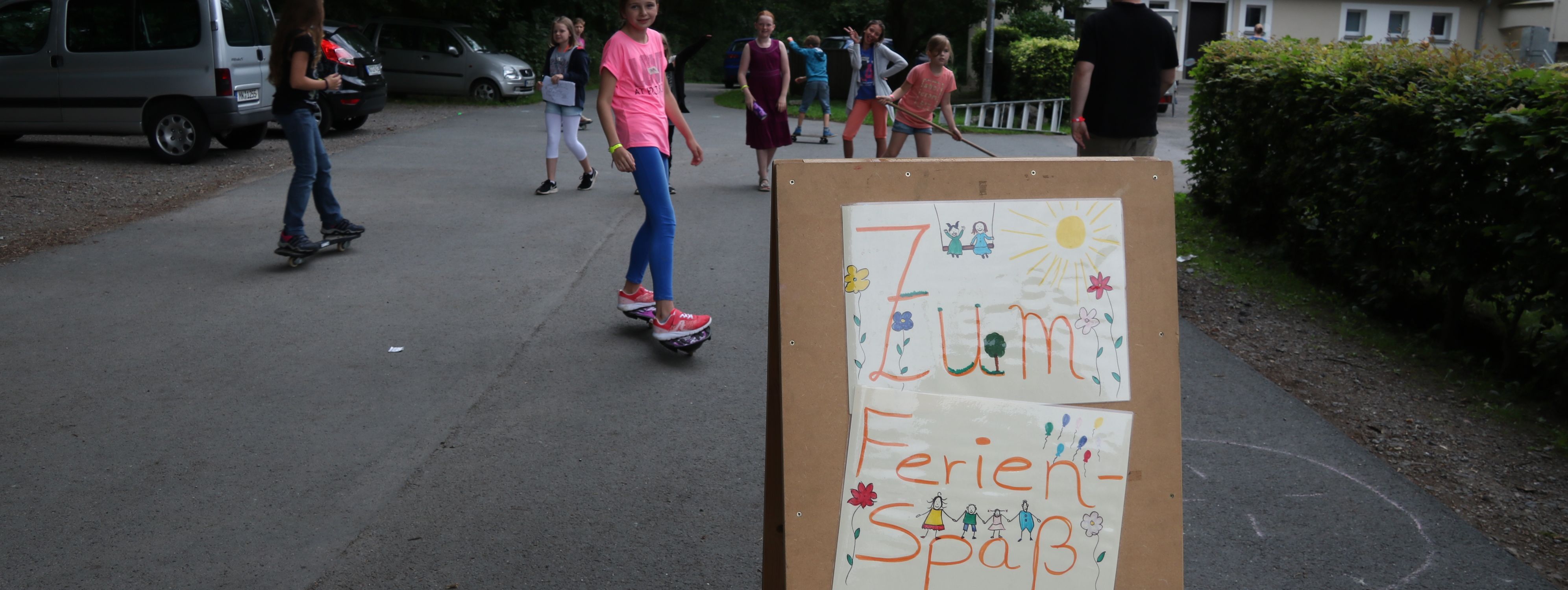 Kinder spielen vor der Jugendbildungsstätte Kluse. Im Vordergrund steht ein Holzschild, das auf den Ferienspaß hinweißt.