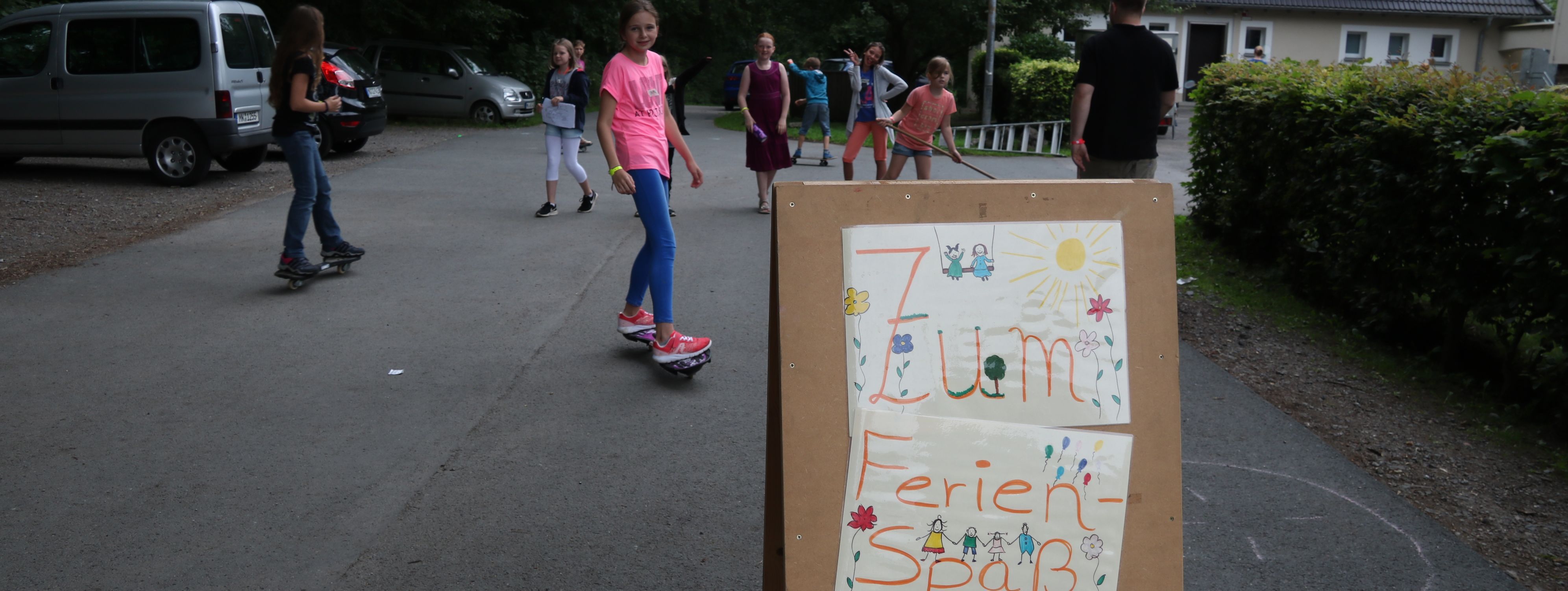 Kinder spielen vor der Jugendbildungsstätte Kluse. Im Vordergrund steht ein Holzschild, das auf den Ferienspaß hinweißt.