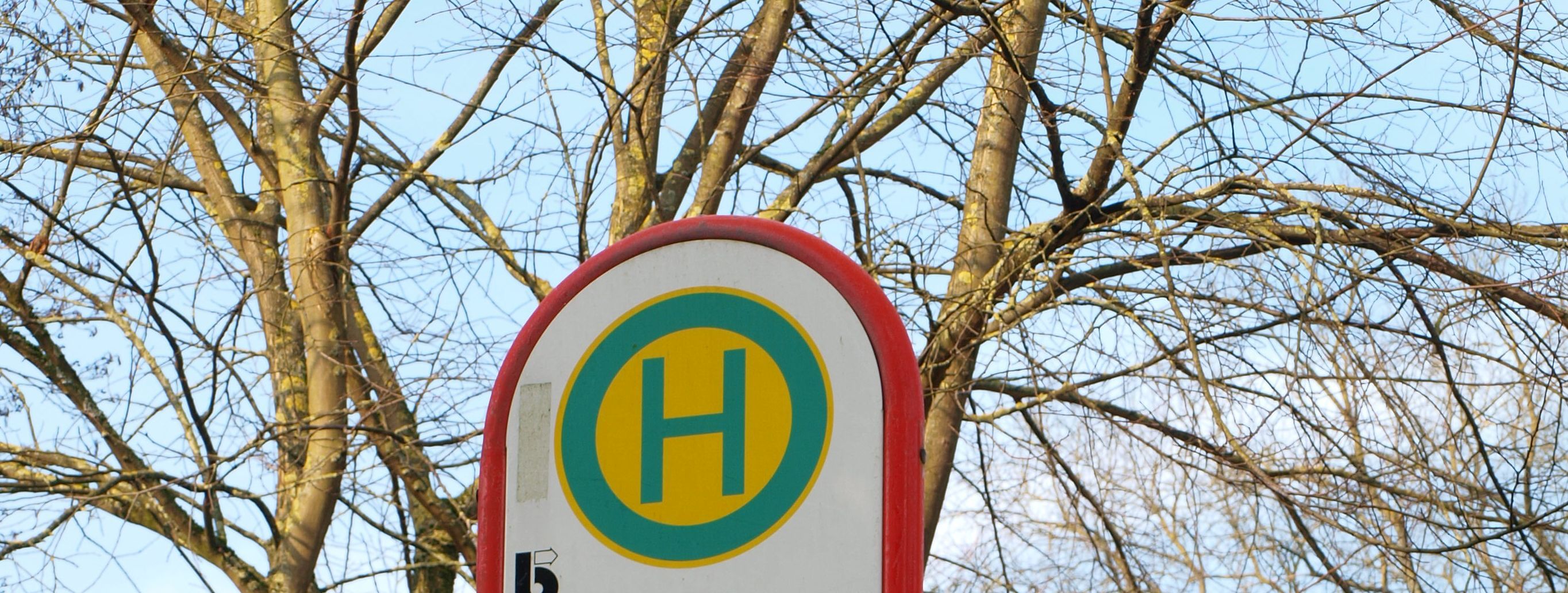 Das Bild zeigt das Schild der Bushaltestelle Menden Bahnhof. Im Hintergrund sind eine Zeitanzeige und Bäume.