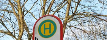 Das Bild zeigt das Schild der Bushaltestelle Menden Bahnhof. Im Hintergrund sind eine Zeitanzeige und Bäume.