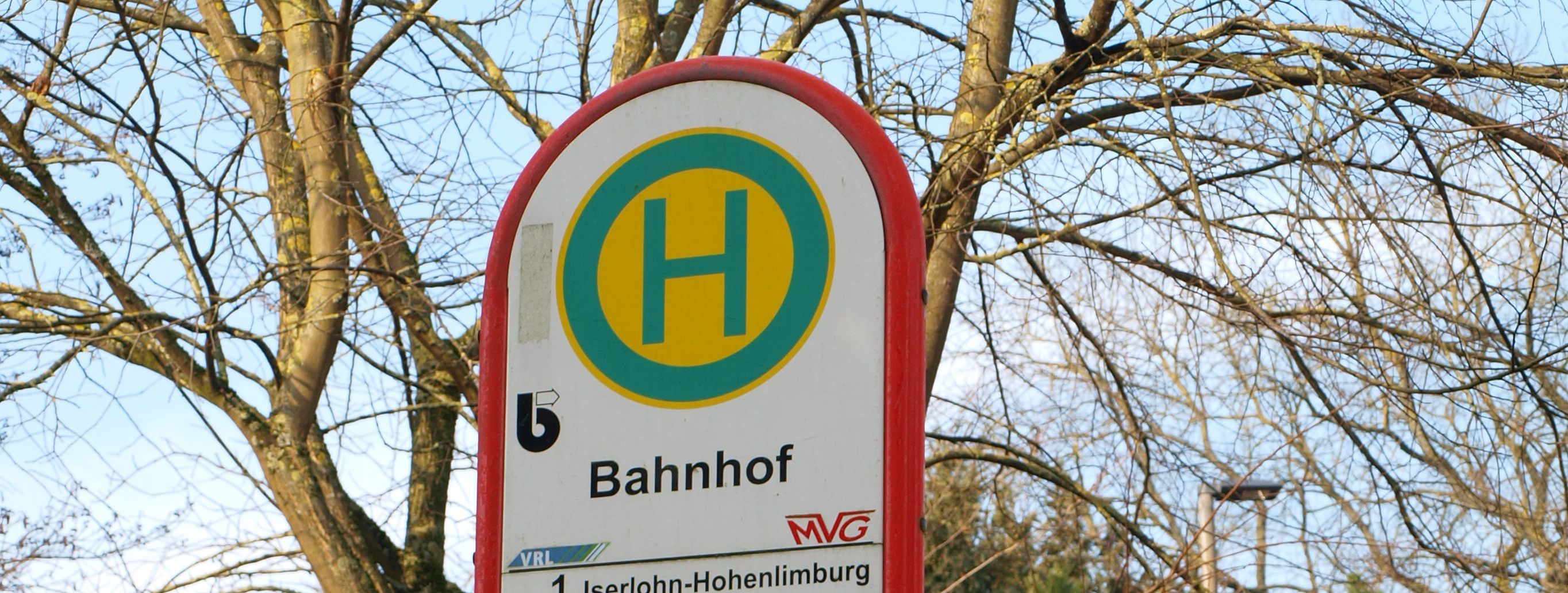 Das Bild zeigt das Schild der Bushaltestelle Menden Bahnhof. Im Hintergrund sind eine Zeitanzeige und Bäume.