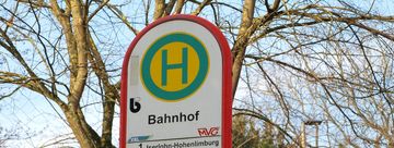 Das Bild zeigt das Schild der Bushaltestelle Menden Bahnhof. Im Hintergrund sind eine Zeitanzeige und Bäume.