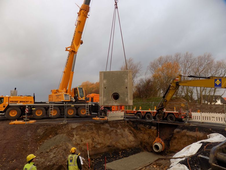 Für den geregelten Ablauf des Regenwassers aus dem Regenrückhaltebecken (RRB) des ersten Bauabschnittes ist ein besonderes Bauwerk erforderlich, das als kompletter Fertigteilschacht mittels Tieflader geliefert und mit einem Kran auf den vorgesehenen Platz versetzt wurde. Das Bauwerk ist in etwa 3,70 m lang, 2,70 m breit und ohne Geländer 2,80 m hoch. Es besteht komplett aus Stahlbeton mit ca. 35 cm dicken Wänden und ist oben offen. Der Kran hat den Schacht vom Tieflader abgehoben und setzt ihn in die Baugrube auf das vorbereitete Fundament (Foto: Stadtentwässerung).