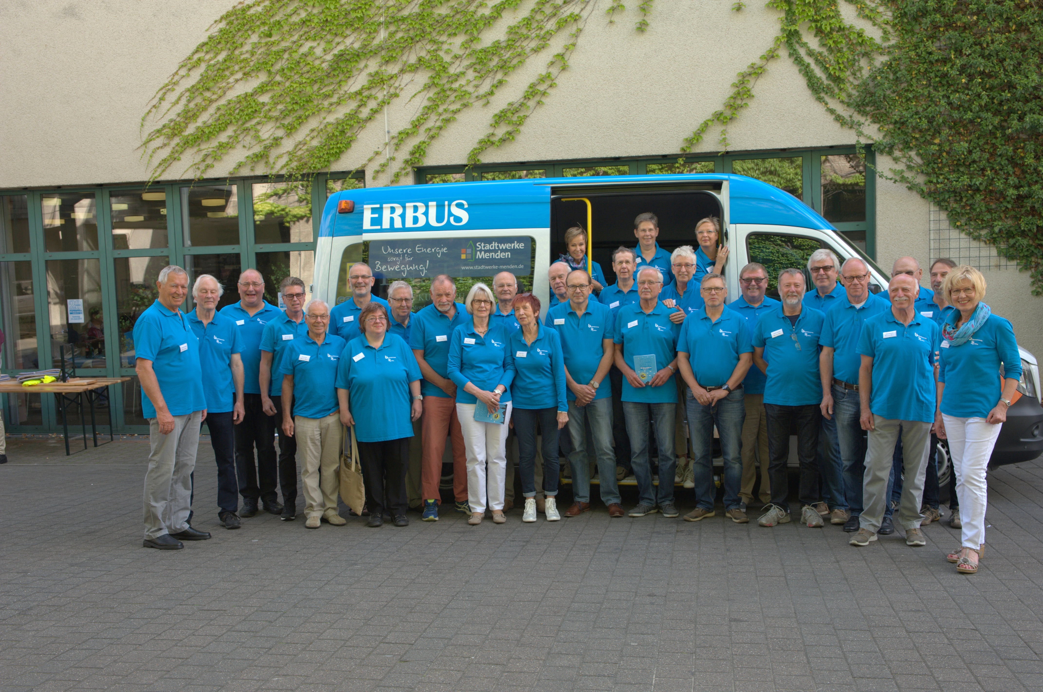 Die Fahrer des Bürgerbusses und die Vereinsmitglieder haben sich vor dem Bürgerbus aufgestellt. Alle tragen die gleichen Polo-Hemden in blau.